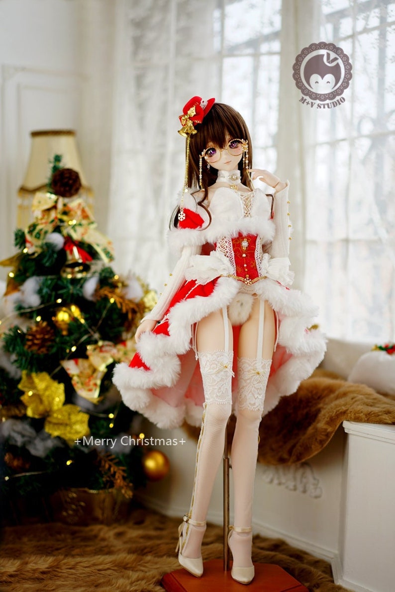 May include: A doll wearing a white and red Christmas dress with a white fur trim. The doll is wearing white stockings and white heels. The doll is standing on a wooden stand. The doll has brown hair and is wearing a red hat with a bell. The doll is holding a white and red ribbon. The doll is standing in front of a Christmas tree.