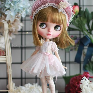 May include: A doll wearing a pink and white dress with a pink and white hat. The doll has blonde hair and blue eyes. The doll is standing on a wooden floor with flowers in the background.
