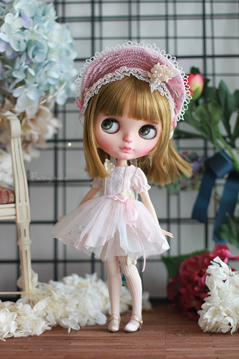 May include: A doll wearing a pink and white dress with a pink and white hat. The doll has blonde hair and blue eyes. The doll is standing on a wooden floor with flowers in the background.