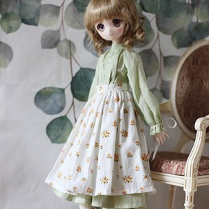 May include: A doll wearing a green blouse and a white skirt with a floral pattern. The doll has long brown hair and is standing in front of a green and white background.