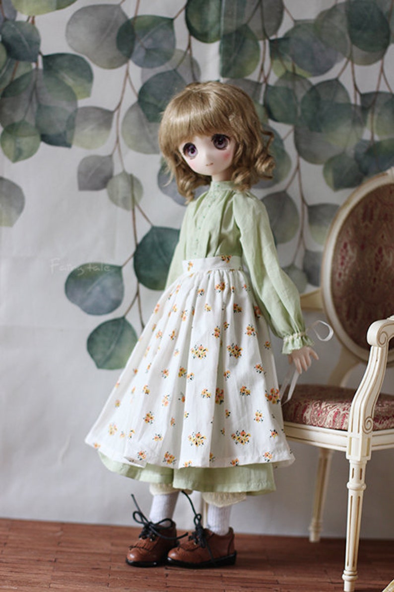 May include: A doll wearing a green blouse and a white skirt with a floral pattern. The doll has long brown hair and is standing in front of a green and white background.