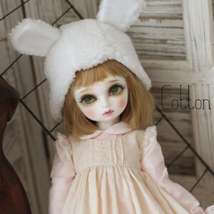 May include: A doll with blonde hair wearing a white fuzzy hat with bunny ears and a pink dress with lace trim. The doll is standing in front of a wooden background.