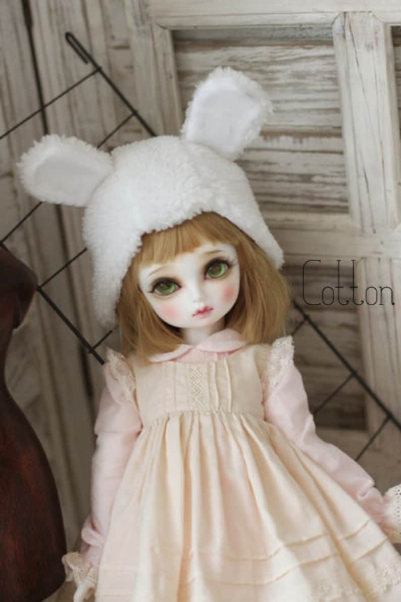 May include: A doll with blonde hair wearing a white fuzzy hat with bunny ears and a pink dress with lace trim. The doll is standing in front of a wooden background.