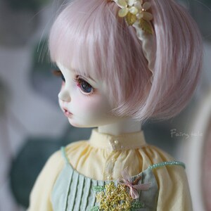 May include: A doll with pink hair wearing a light green dress with yellow flower embroidery and a white headband with yellow flowers.