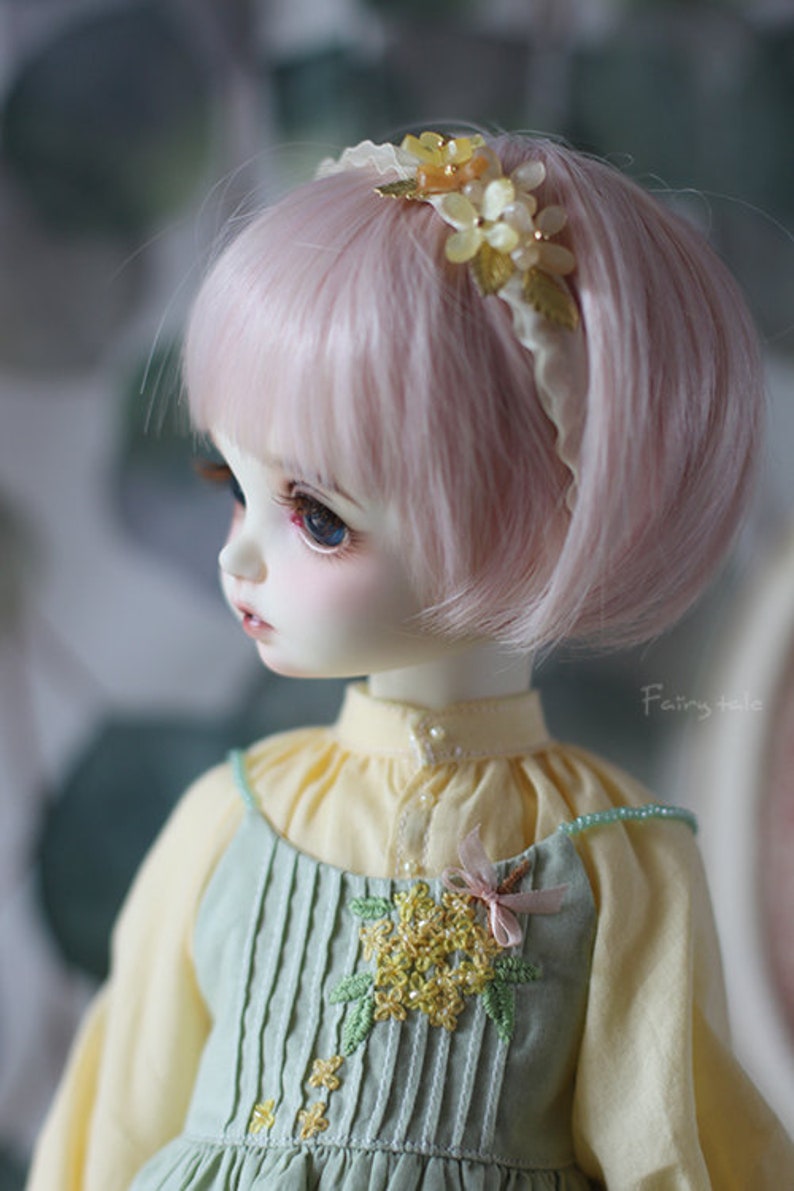 May include: A doll with pink hair wearing a light green dress with yellow flower embroidery and a white headband with yellow flowers.