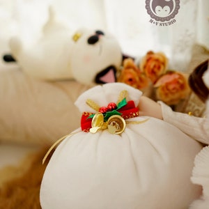 May include: A white fabric sack with a red ribbon and gold bells, decorated with a green sprig of holly and red berries. The sack is sitting on a brown furry surface. +Merry Christmas+ is written below the sack.