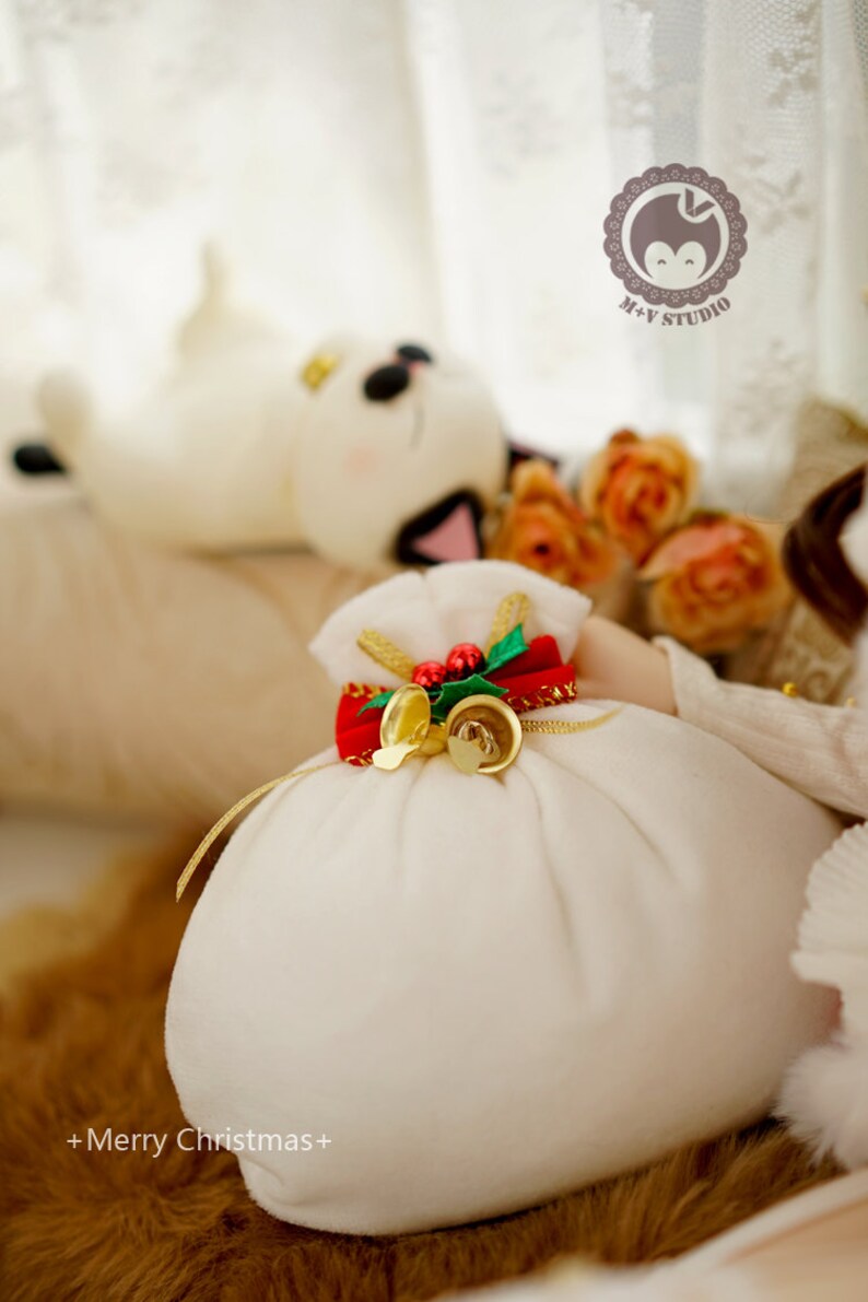 May include: A white fabric sack with a red ribbon and gold bells, decorated with a green sprig of holly and red berries. The sack is sitting on a brown furry surface. +Merry Christmas+ is written below the sack.