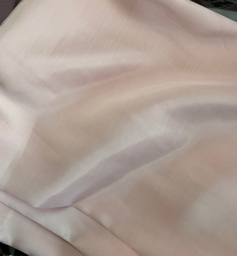 May include: A pale pink fabric with a smooth, silky texture. The fabric is folded and draped, showing its soft, flowing nature.