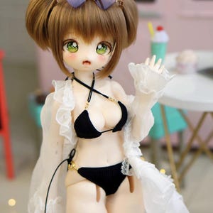 May include: A doll with brown hair styled in two buns, wearing heart-shaped sunglasses. The doll is dressed in a black bikini with gold accents and a white, ruffled cover-up. The doll has large green eyes and rosy cheeks.