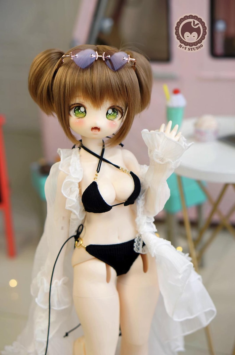 May include: A doll with brown hair styled in two buns, wearing heart-shaped sunglasses. The doll is dressed in a black bikini with gold accents and a white, ruffled cover-up. The doll has large green eyes and rosy cheeks.