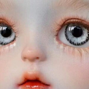 Op de afbeelding: Close-up van het gezicht van een pop met grote, witte ogen met zwarte pupillen en een gedetailleerde iris. De pop heeft een lichte huid en een kleine, roze mond.
