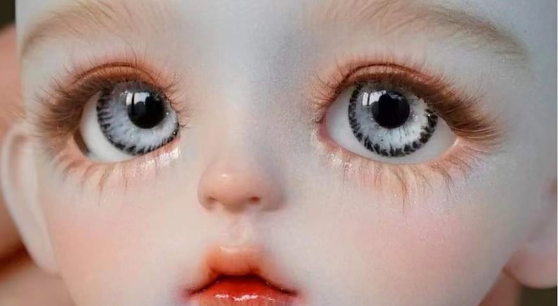 Op de afbeelding: Close-up van het gezicht van een pop met grote, witte ogen met zwarte pupillen en een gedetailleerde iris. De pop heeft een lichte huid en een kleine, roze mond.