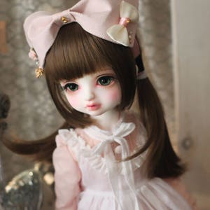 May include: A doll with long brown hair wearing a pink dress and a pink bow with white accents.