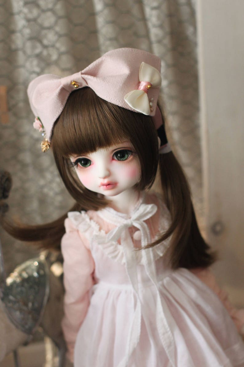 May include: A doll with long brown hair wearing a pink dress and a pink bow with white accents.