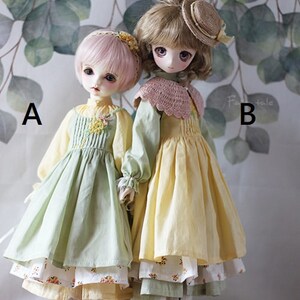 May include: Two dolls wearing dresses with floral patterns. The doll on the left is wearing a green dress with a yellow underskirt and the doll on the right is wearing a yellow dress with a green underskirt. Both dolls are wearing brown shoes.
