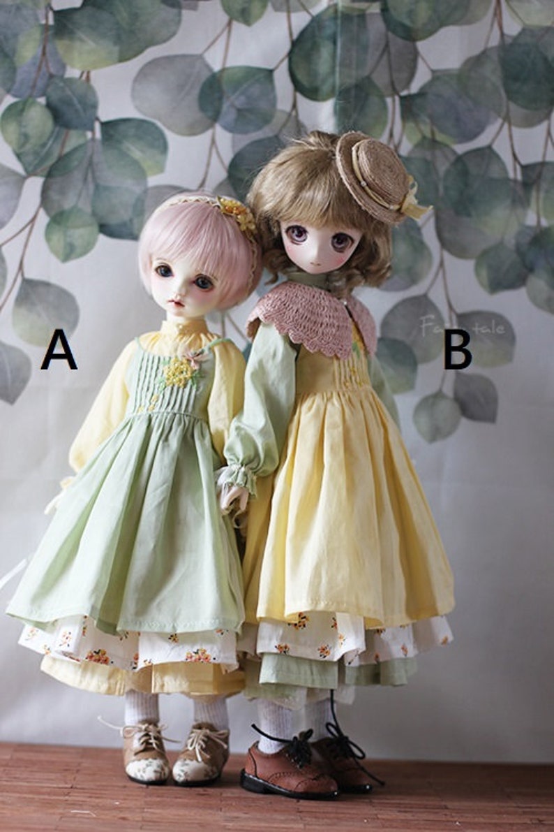 May include: Two dolls wearing dresses with floral patterns. The doll on the left is wearing a green dress with a yellow underskirt and the doll on the right is wearing a yellow dress with a green underskirt. Both dolls are wearing brown shoes.