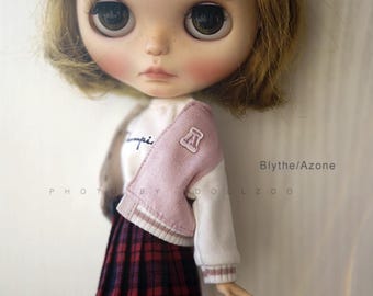 May include: A Blythe doll with large eyes and freckles, wearing a white and pink letterman jacket over a white shirt and a plaid skirt. The jacket has a patch with the letter "A". The doll is a collectible item.
