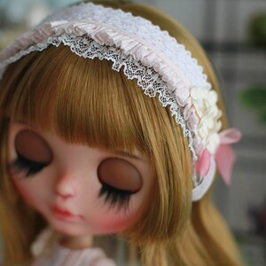 May include: A doll with blonde hair and closed eyes wears a white headband with lace and pink fabric accents. The headband has a flower embellishment.