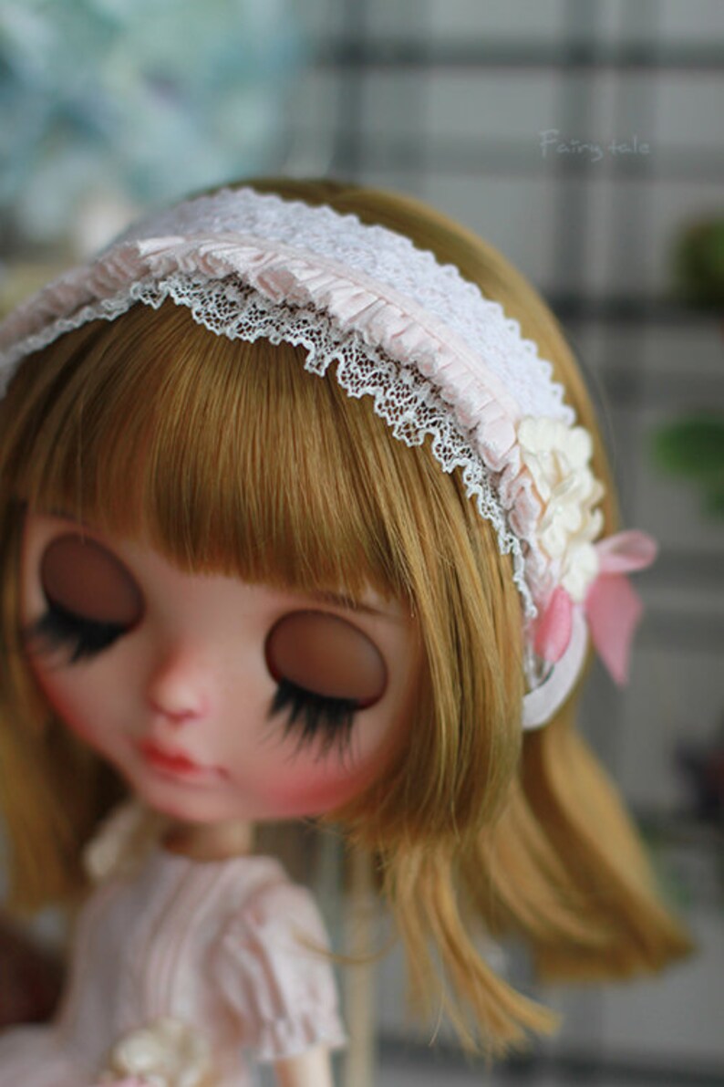 May include: A doll with blonde hair and closed eyes wears a white headband with lace and pink fabric accents. The headband has a flower embellishment.