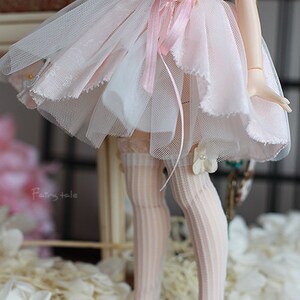 May include: A doll wearing a pink and white tulle dress with a pink ribbon and a white flower. The doll is wearing white stockings and pink shoes. The doll is standing on a white surface with white flowers.