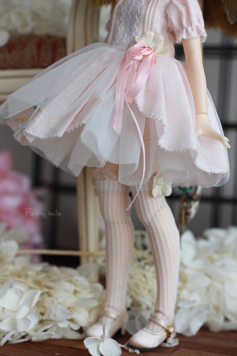 May include: A doll wearing a pink and white tulle dress with a pink ribbon and a white flower. The doll is wearing white stockings and pink shoes. The doll is standing on a white surface with white flowers.