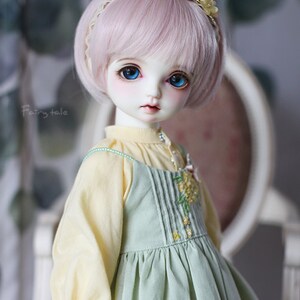 May include: A doll with pink hair and blue eyes wearing a light green dress with a yellow blouse and a flower headband.