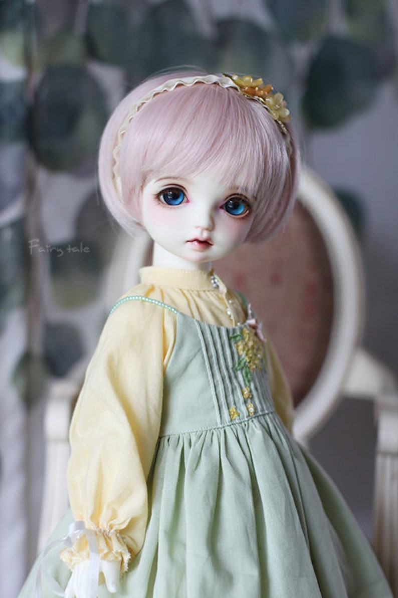 May include: A doll with pink hair and blue eyes wearing a light green dress with a yellow blouse and a flower headband.