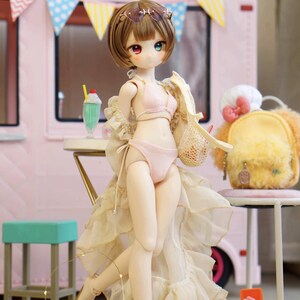 May include: A doll in a pink bikini and a sheer, ruffled cover-up stands in front of a pink food truck. The doll has brown hair, sunglasses, and mismatched eyes. A yellow backpack and a green milkshake are also visible.