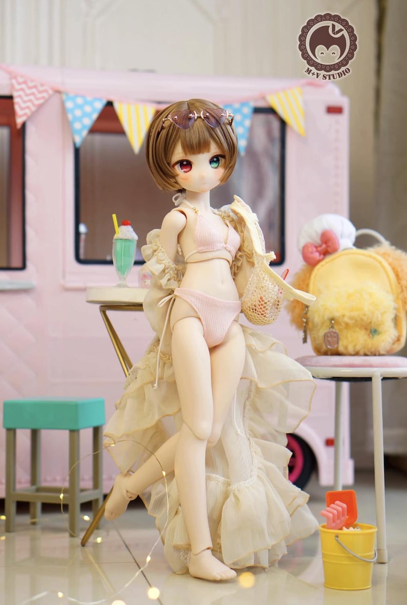 May include: A doll in a pink bikini and a sheer, ruffled cover-up stands in front of a pink food truck. The doll has brown hair, sunglasses, and mismatched eyes. A yellow backpack and a green milkshake are also visible.