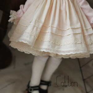May include: A white cotton dress with lace trim and a pink bodice. The dress has a gathered skirt and is worn by a doll with white tights and black shoes.