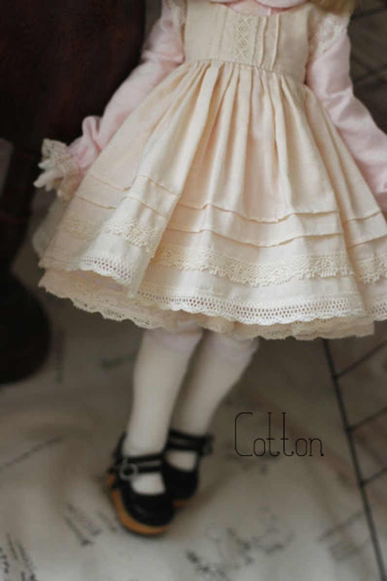 May include: A white cotton dress with lace trim and a pink bodice. The dress has a gathered skirt and is worn by a doll with white tights and black shoes.