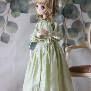 May include: A doll wearing a light green dress with white ribbon accents and brown shoes. The doll has blonde hair and brown eyes. The doll is standing in front of a green and white background.