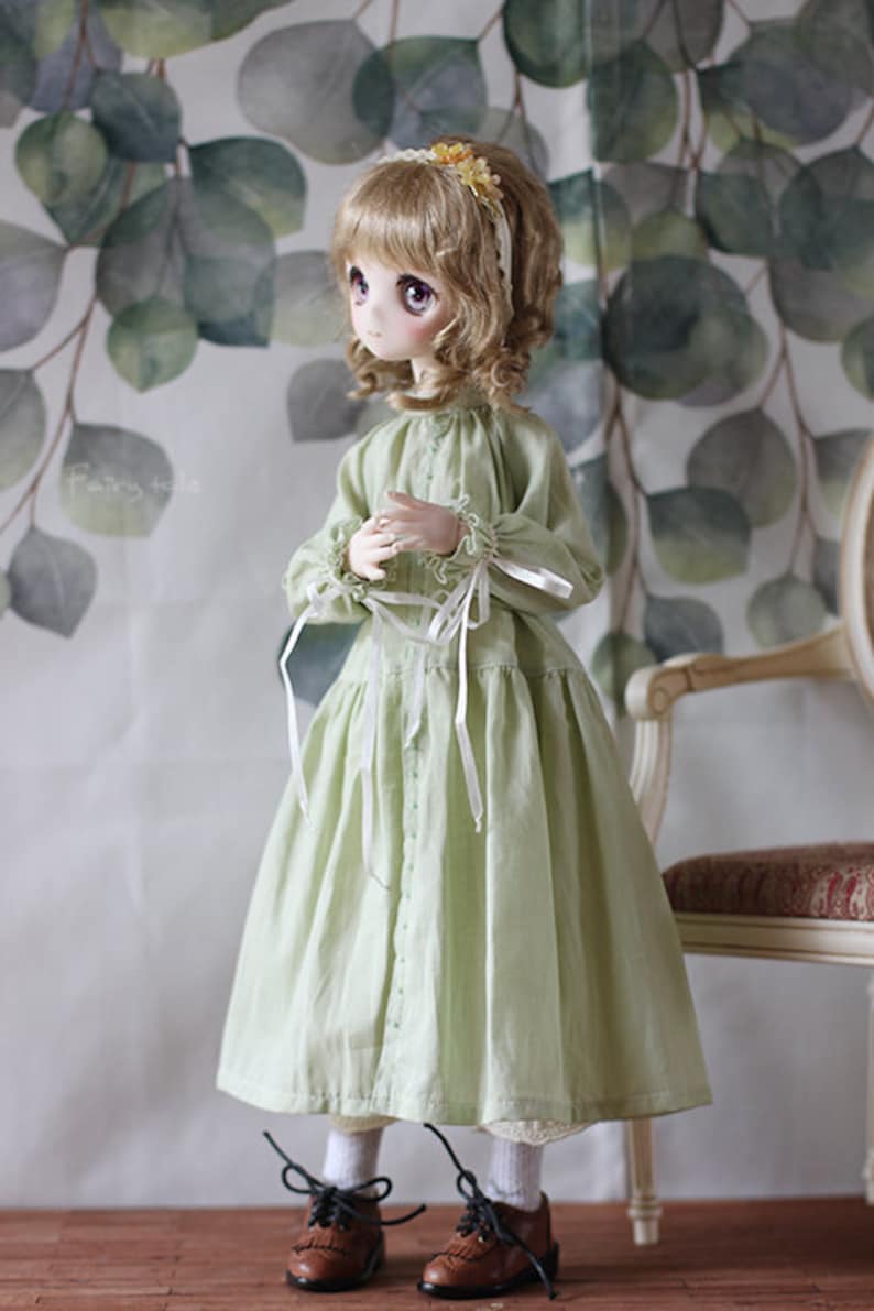 May include: A doll wearing a light green dress with white ribbon accents and brown shoes. The doll has blonde hair and brown eyes. The doll is standing in front of a green and white background.