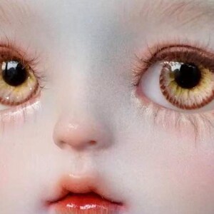 Op de afbeelding: Close-up van het gezicht van een pop met grote, ronde, geel-gouden ogen met bruine iris en donkere pupillen. De pop heeft een bleke huid en een kleine, roze mond.