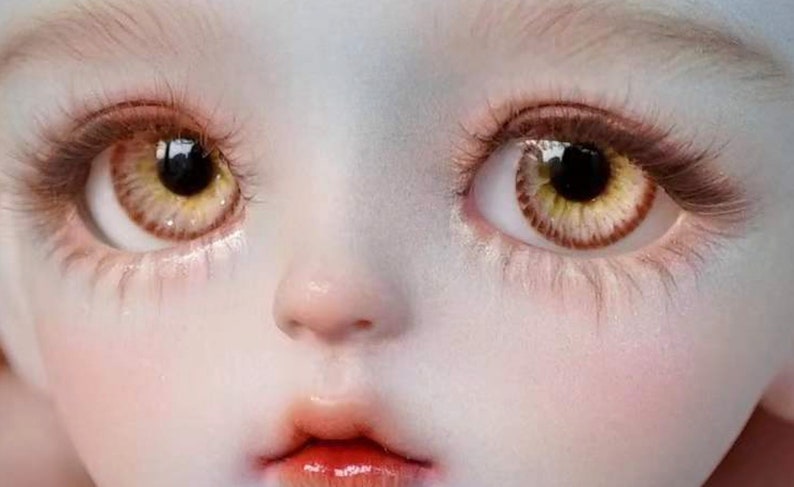 Op de afbeelding: Close-up van het gezicht van een pop met grote, ronde, geel-gouden ogen met bruine iris en donkere pupillen. De pop heeft een bleke huid en een kleine, roze mond.