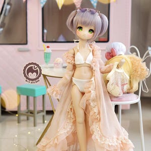 May include: A doll with lavender hair and green eyes, wearing a white bikini and a light pink robe with lace trim. The doll is standing in front of a pink food truck with a banner of colorful flags. A small table has a milkshake and ice cream.