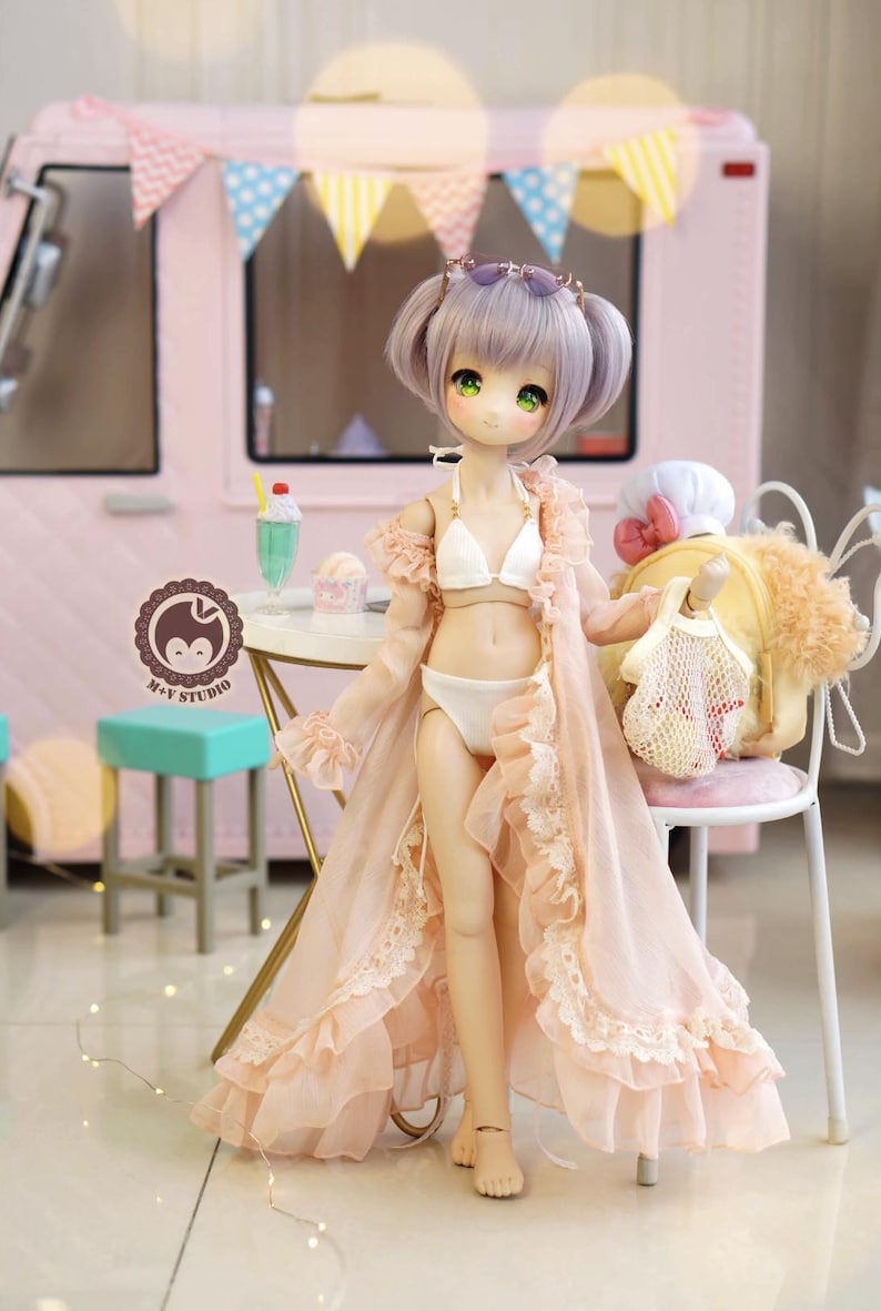 May include: A doll with lavender hair and green eyes, wearing a white bikini and a light pink robe with lace trim. The doll is standing in front of a pink food truck with a banner of colorful flags. A small table has a milkshake and ice cream.