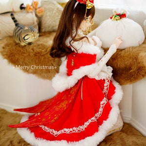 May include: A doll wearing a red and white dress with a white fur trim, a red hat with gold accents, and white shoes. The doll is holding a white bag with a red bow and gold bells. The doll is kneeling on a brown fur rug. +Merry Christmas+ is written on the rug.