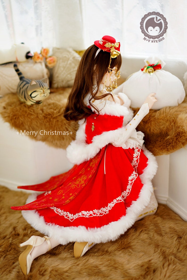 May include: A doll wearing a red and white dress with a white fur trim, a red hat with gold accents, and white shoes. The doll is holding a white bag with a red bow and gold bells. The doll is kneeling on a brown fur rug. +Merry Christmas+ is written on the rug.