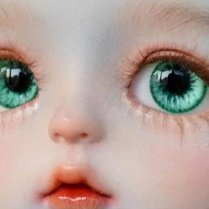 Op de afbeelding: Close-up van het gezicht van een pop met groene ogen. De pop heeft een lichte huidskleur en een kleine, delicate neus. De ogen zijn groot en rond, met een donkere iris en een groene buitenring.