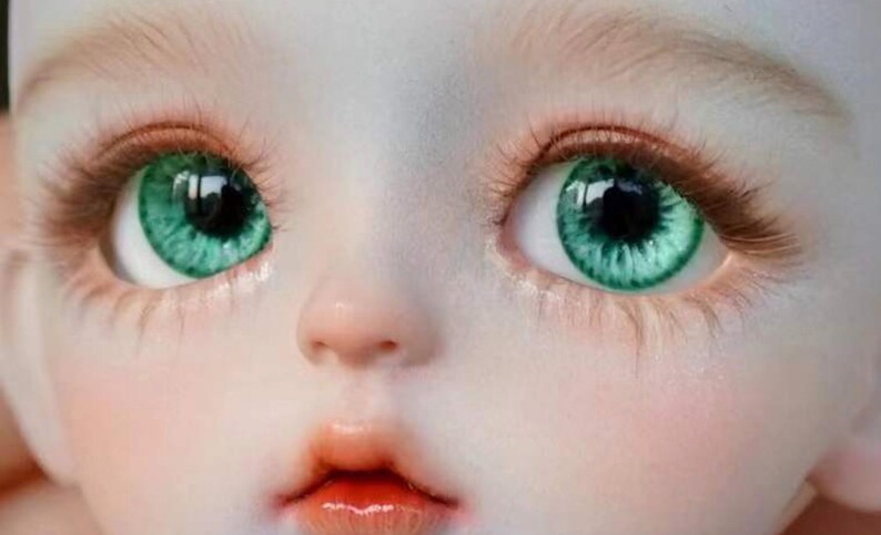 Op de afbeelding: Close-up van het gezicht van een pop met groene ogen. De pop heeft een lichte huidskleur en een kleine, delicate neus. De ogen zijn groot en rond, met een donkere iris en een groene buitenring.