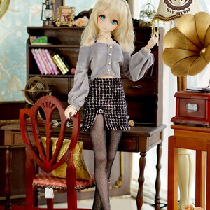 May include: A doll with blonde hair wearing a grey sweater, a black and white plaid skirt, black tights, and black shoes. The doll is standing in a room with a wooden desk, a chair, and a lamp with a brown and white lampshade. The lampshade has the text "M+V STUDIO" on it.