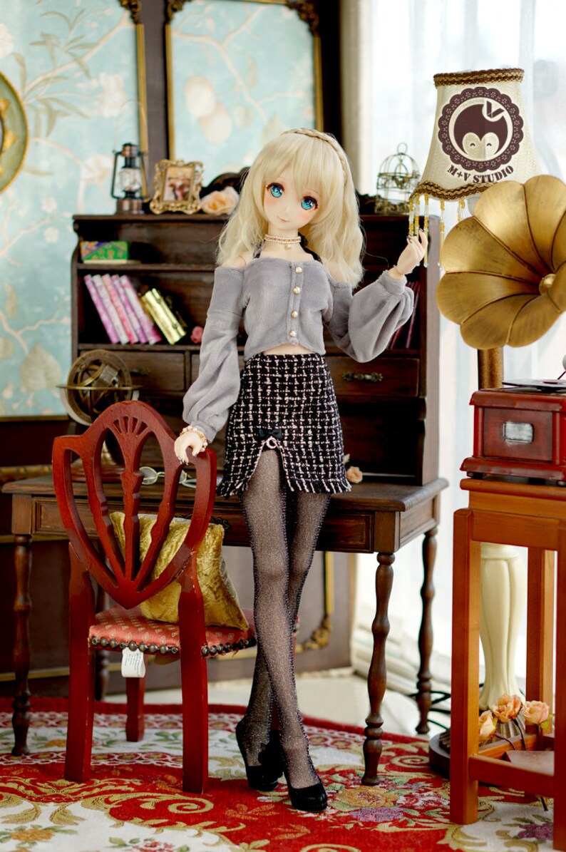 May include: A doll with blonde hair wearing a grey sweater, a black and white plaid skirt, black tights, and black shoes. The doll is standing in a room with a wooden desk, a chair, and a lamp with a brown and white lampshade. The lampshade has the text "M+V STUDIO" on it.