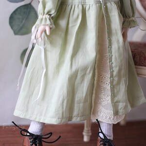 May include: A doll wearing a light green dress with white lace trim and brown leather shoes with black laces.