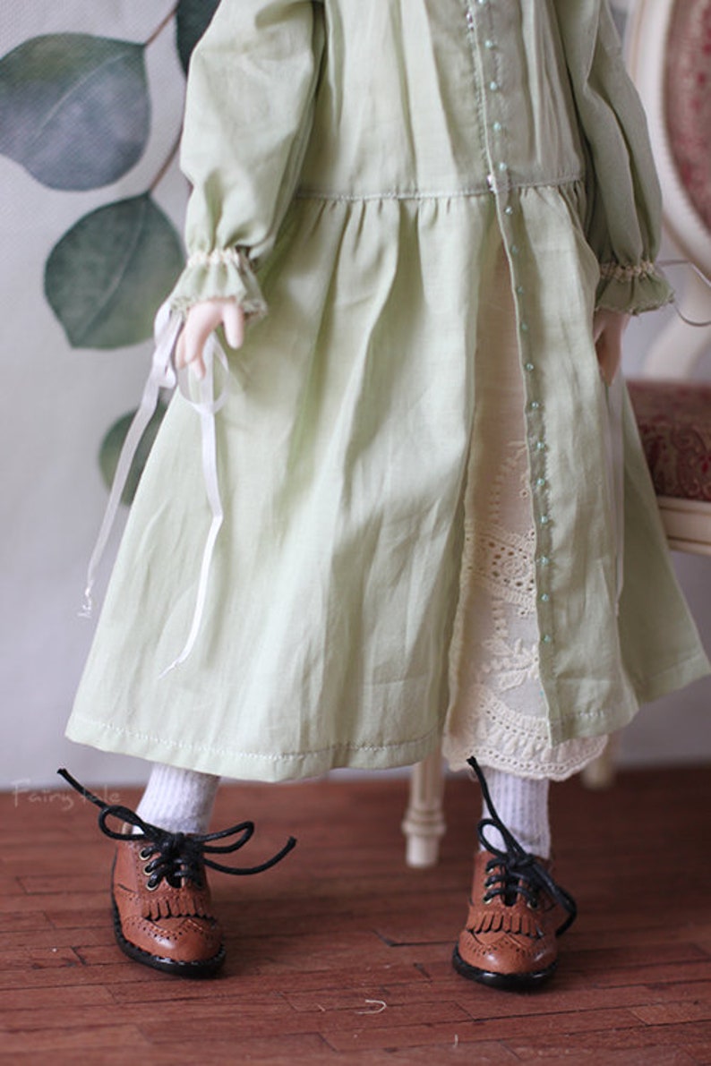 May include: A doll wearing a light green dress with white lace trim and brown leather shoes with black laces.