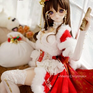 May include: A doll wearing a red and white Christmas dress with a white fur trim, a red hat with gold accents, and gold-rimmed glasses. The doll is holding a rolled-up scroll in one hand and has a white furry bag with gold accents next to her.
