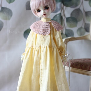 May include: A doll wearing a yellow dress with a pink crocheted collar and brown shoes. The doll has pink hair and blue eyes. The doll is standing in front of a chair with a floral patterned fabric.