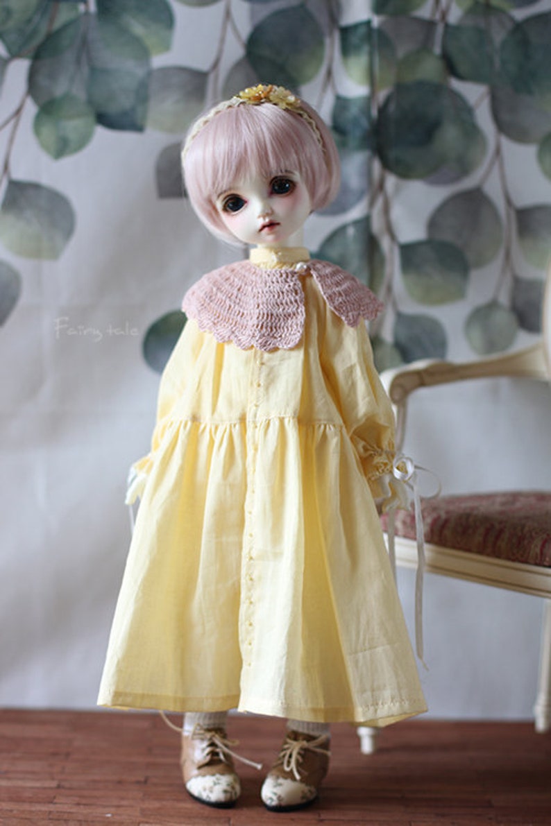 May include: A doll wearing a yellow dress with a pink crocheted collar and brown shoes. The doll has pink hair and blue eyes. The doll is standing in front of a chair with a floral patterned fabric.