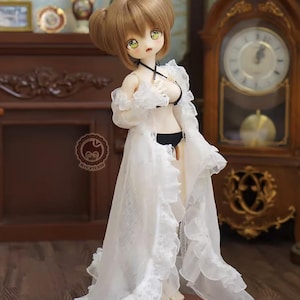 May include: A doll with brown hair styled in two buns, wearing a black bikini and a long, white, ruffled robe. The doll is standing on a wooden base. The background includes a wooden clock and furniture.
