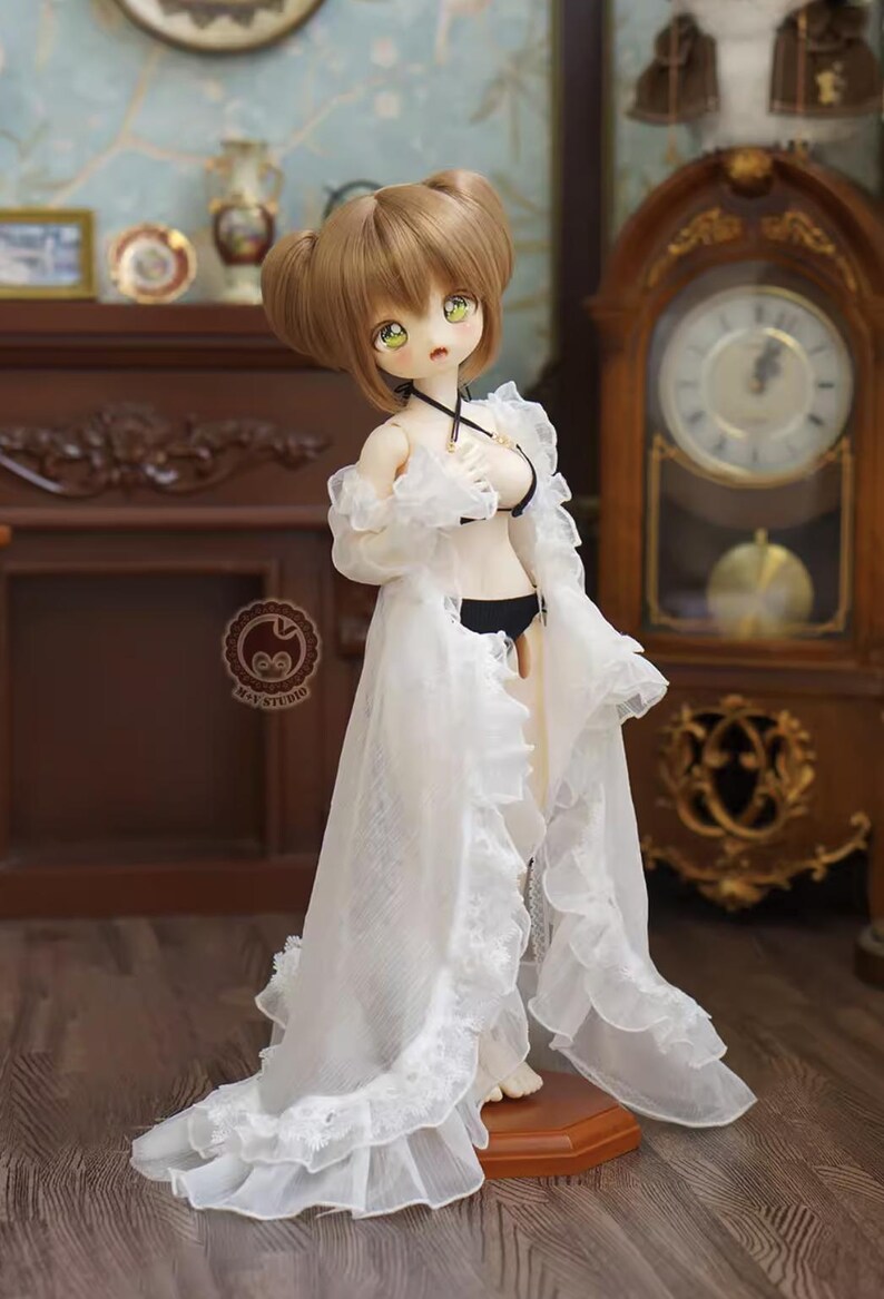 May include: A doll with brown hair styled in two buns, wearing a black bikini and a long, white, ruffled robe. The doll is standing on a wooden base. The background includes a wooden clock and furniture.